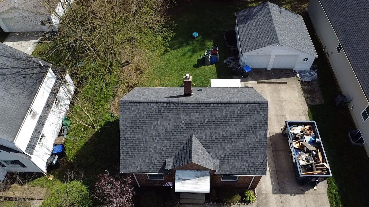 Aerial view of a house with a gray roof, a driveway, a detached garage, and a dumpster filled with debris next to the driveway.