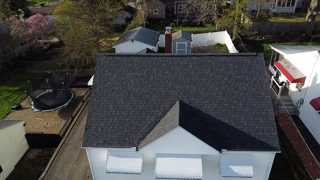 Aerial view of a house with a gray shingle roof, a detached garage, a trampoline in the yard, and neighboring houses visible.