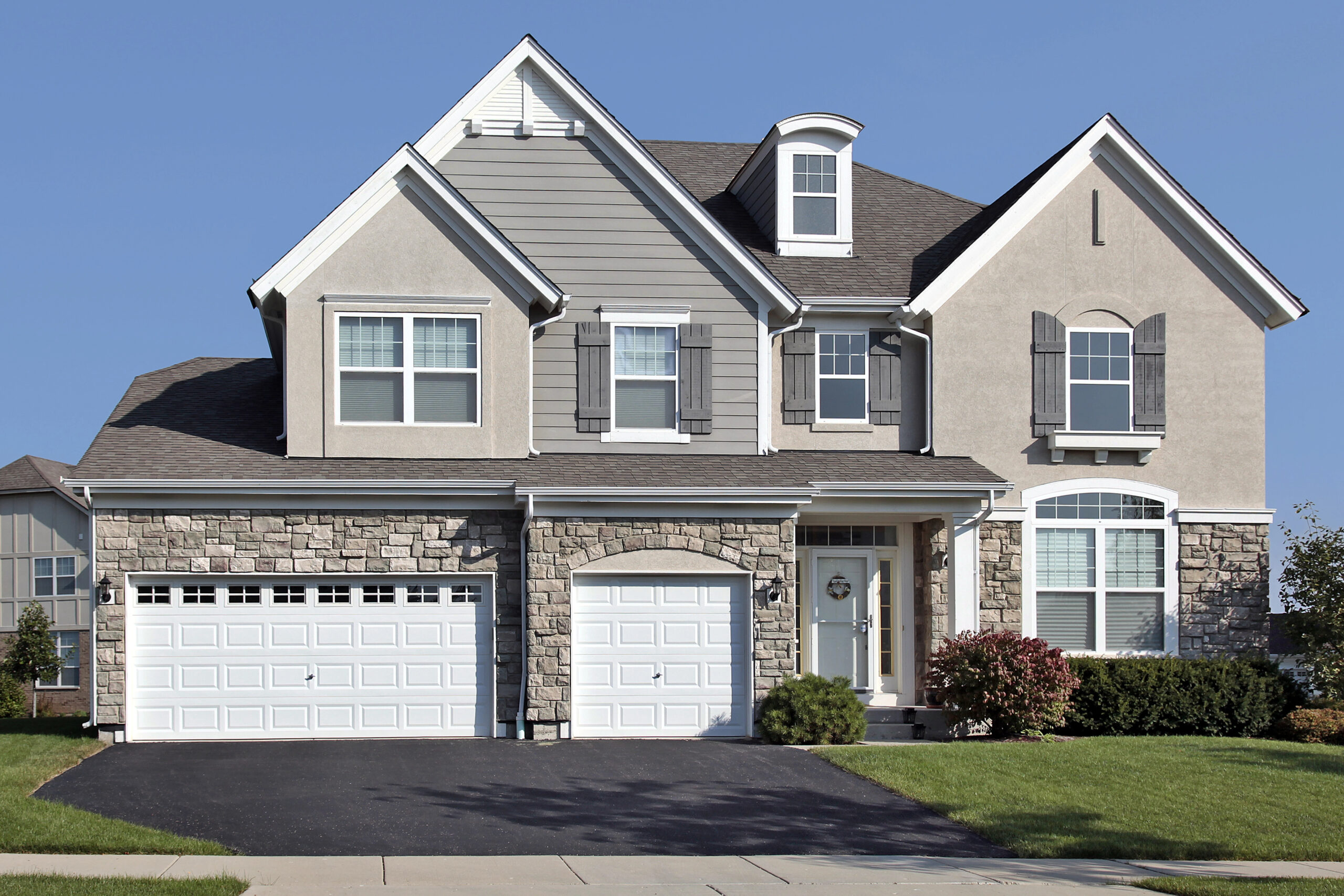 Two-story suburban house with gray siding, stone accents, three white garage doors, shuttered windows, a small front porch, and a neatly trimmed lawn under a clear sky.