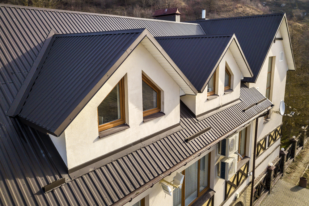 Aerial view of a beige house with a dark metal gable roof, triangular dormer windows, and air conditioning units mounted on the exterior wall.