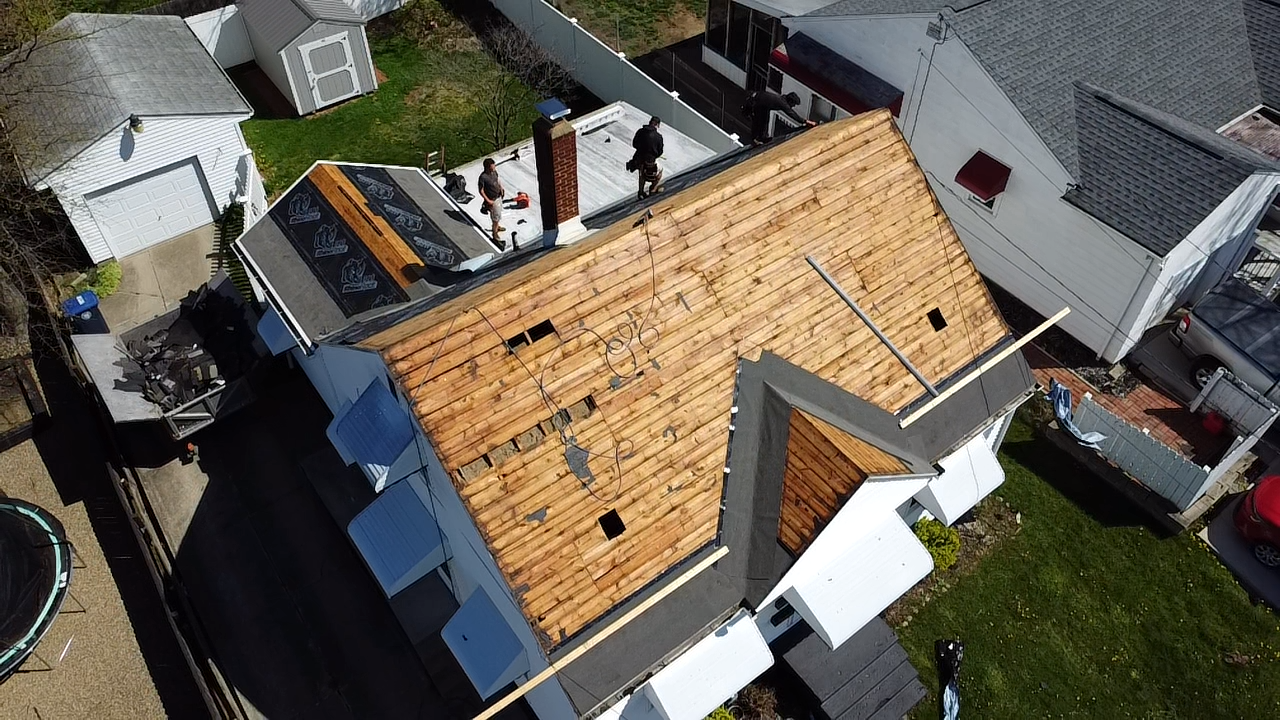 Aerial view of a house roof under repair with exposed wooden decking and construction materials; a worker is present on the roof.
