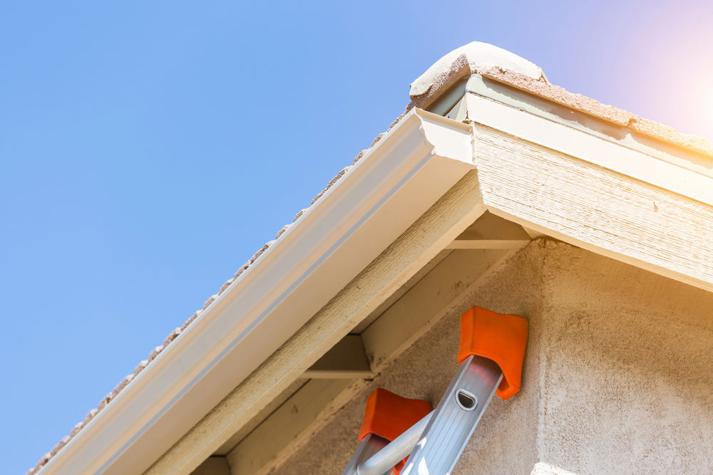 A close-up view of a house roofline with a white gutter, beige siding, and a ladder leaning against the wall under a clear blue sky.