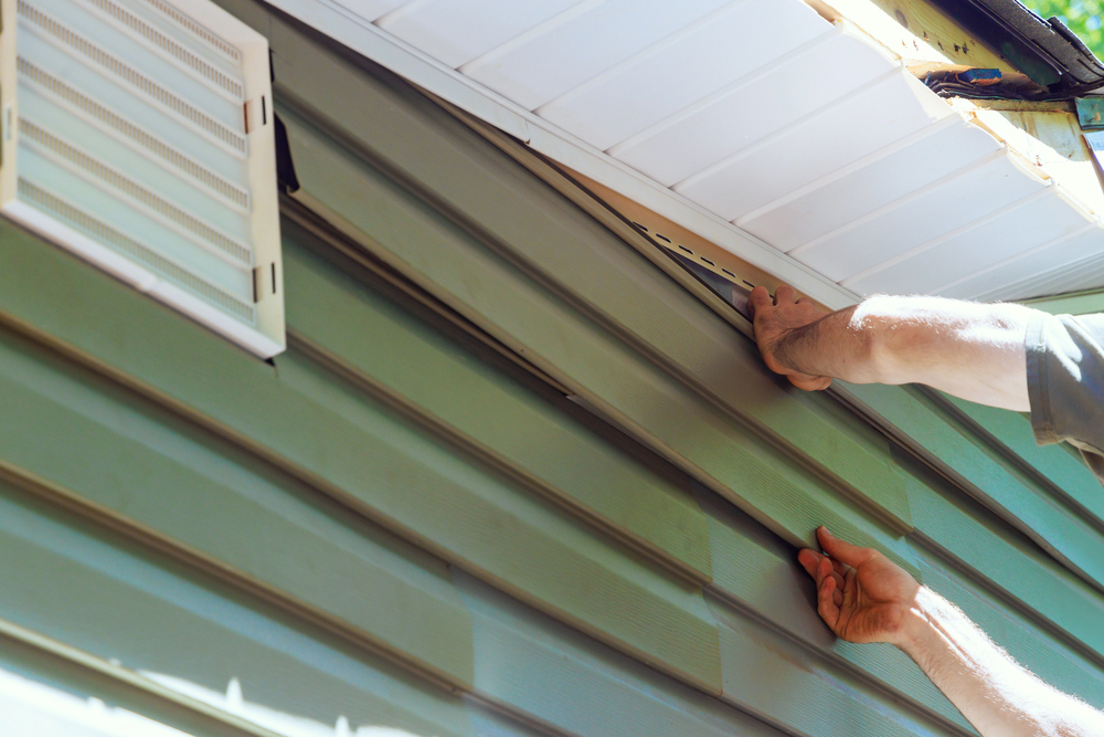 A person installs green vinyl siding panels on the exterior wall of a house near a white vent and soffit.